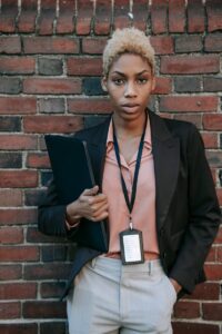 Femme professionnelle aux cheveux courts posant avec assurance contre un mur de briques, tenant un ordinateur portable.